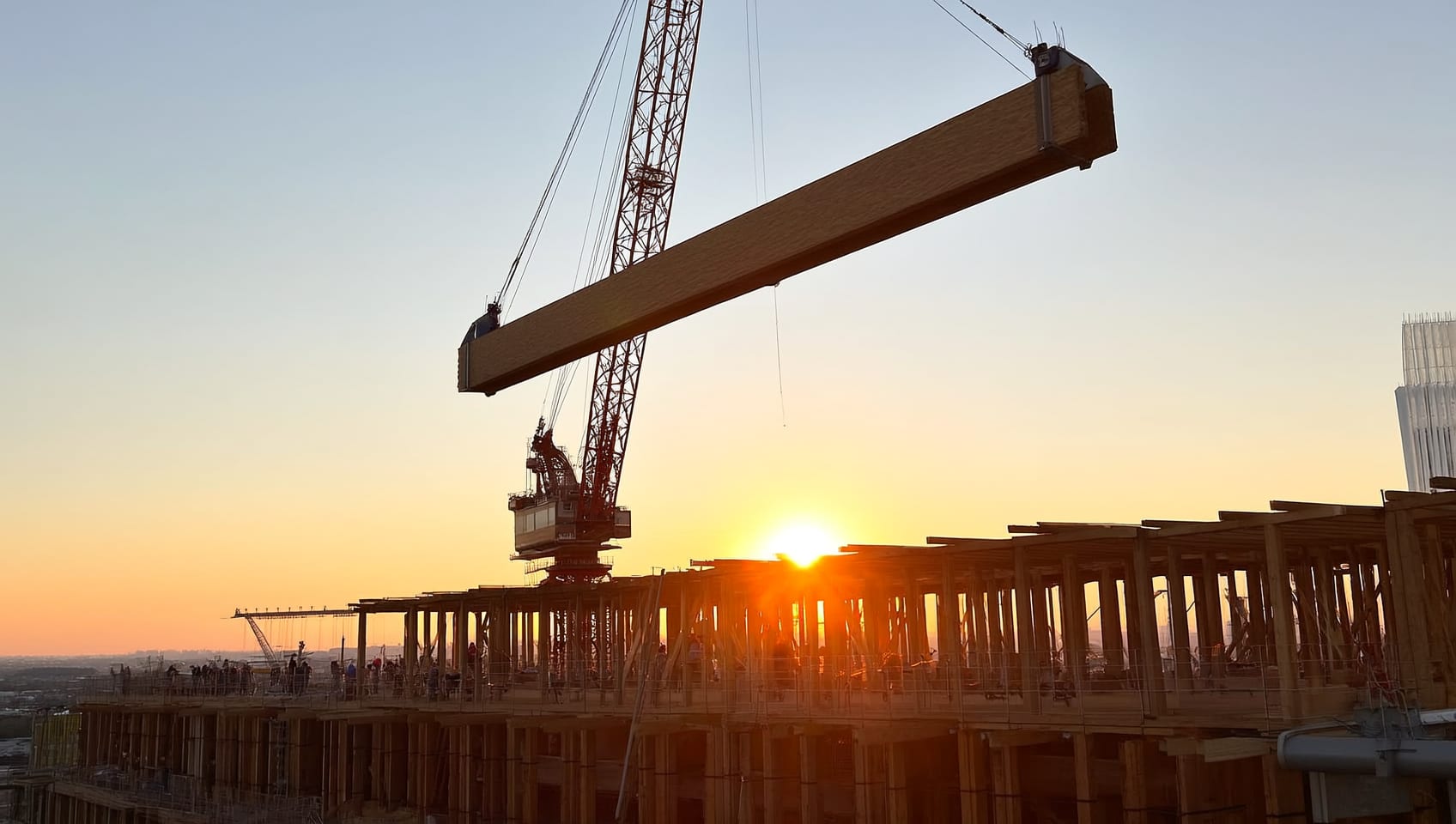 Tower crane lifting a large timber beam over a rising wood-framed structure at golden hour